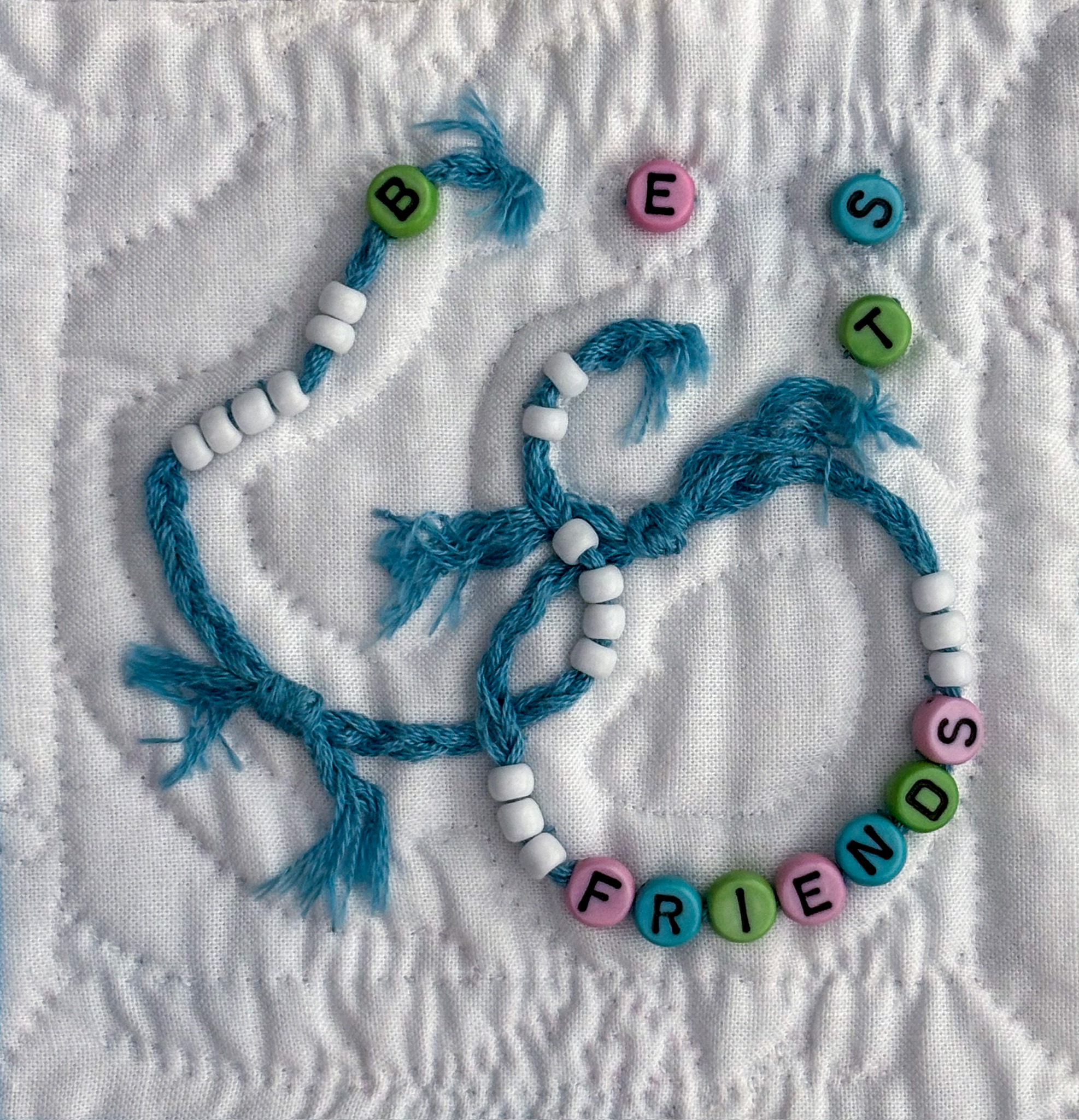 Two interlocking friendship bracelets appliquéd on white cotton. The “BEST” bracelet has burst, scattering the letter beads. The “FRIENDS” bracelet remains intact.
