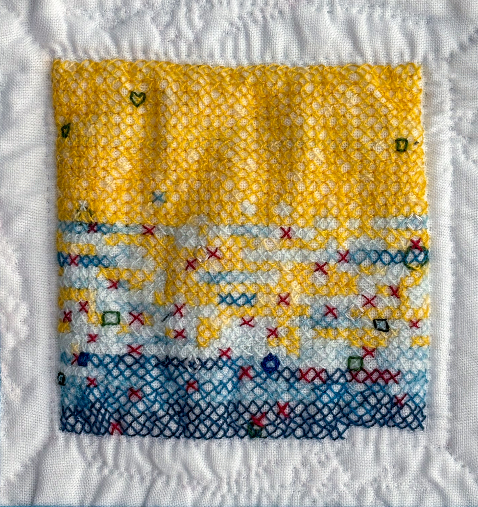 An abstract cross-stitch with a gradient from dark yellow falling into dark blue on white cotton, throughout which shapes such as hearts, stars and squares the size of one full stitch and a few red stitches are scattered.