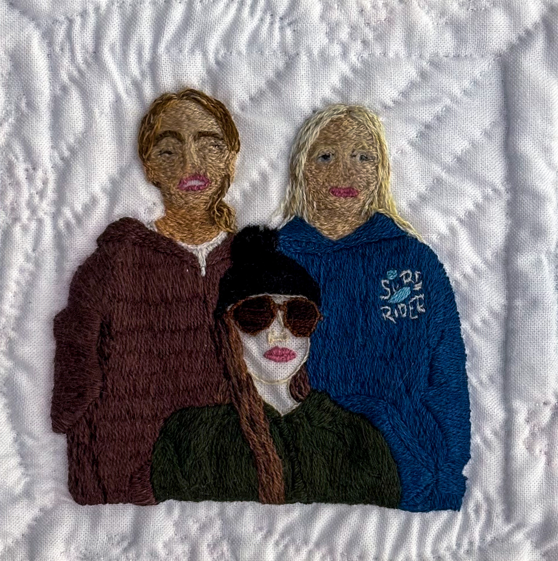 Three young women, embroidered on white cotton. In the foreground, an outlined woman in a toque and sunglasses sits, dressed in a forest green sweater. Just behind her stand two smiling women, densely stitched. On the left, an auburn-haired woman in a brown zip-up grins next to a blonde woman wearing a vibrant blue hoodie with a “Surf Rider” emblem.