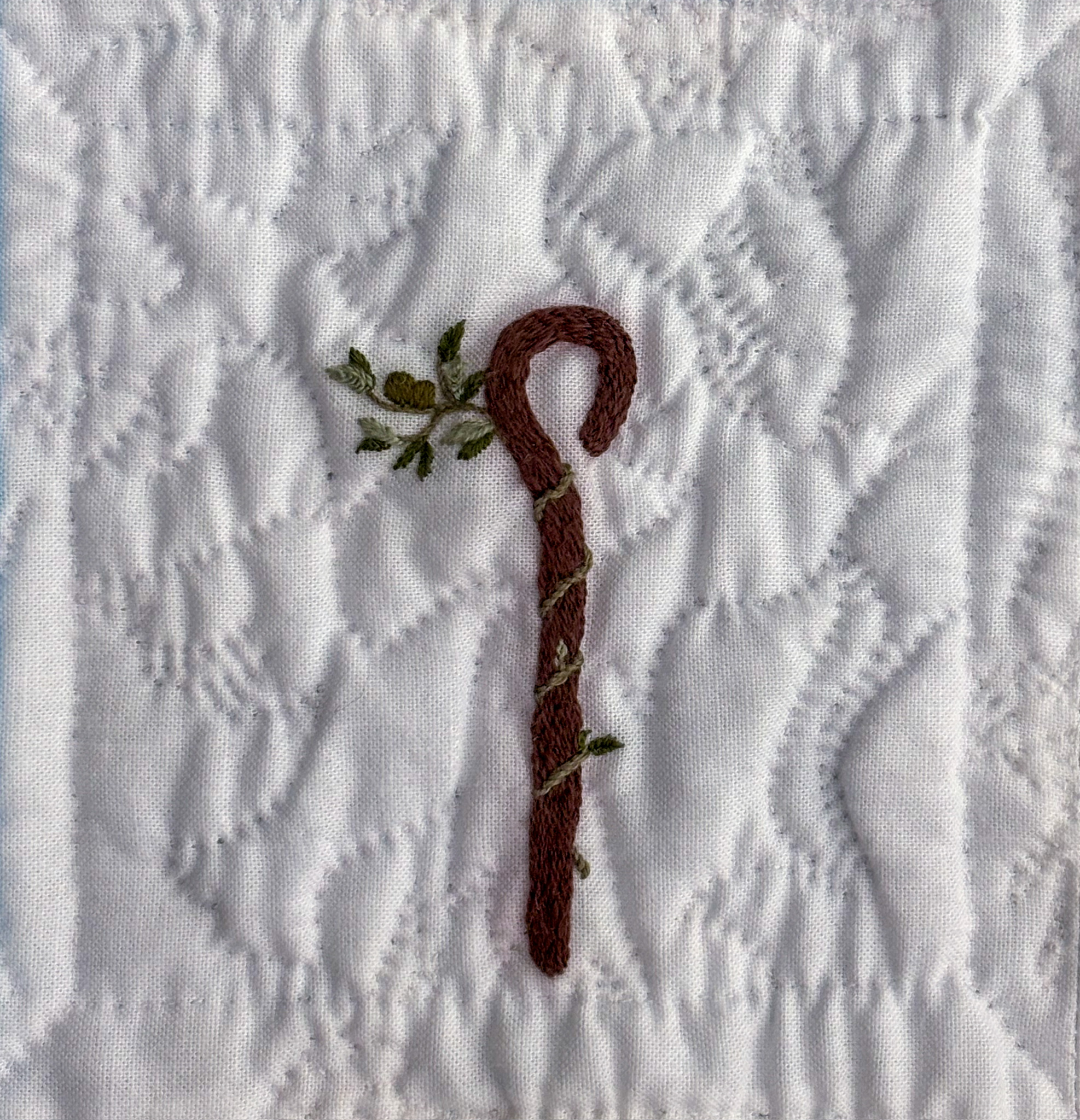 A brown olive branch with three leafy offshoots and a cluster of olives climbing a brown crook embroidered on white cotton.