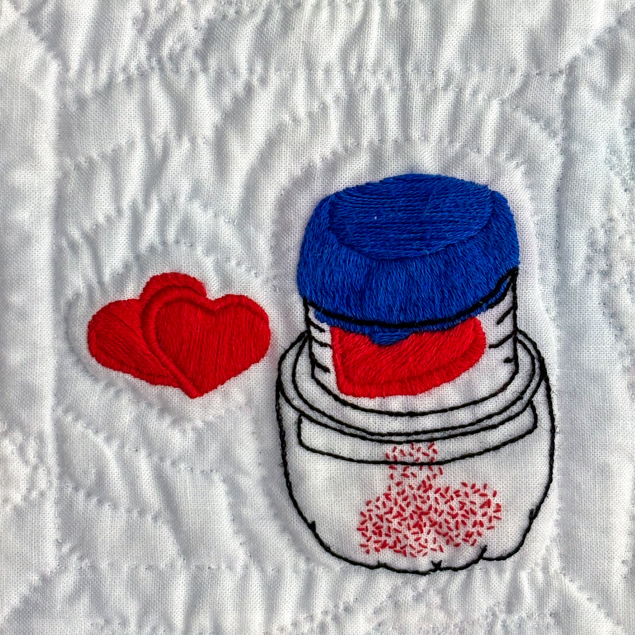 A clear pill crusher with blue lid and two small red heart pills embroidered on white cotton. A similar pill sits in the upper compartment as red particles fall to the lower compartment.