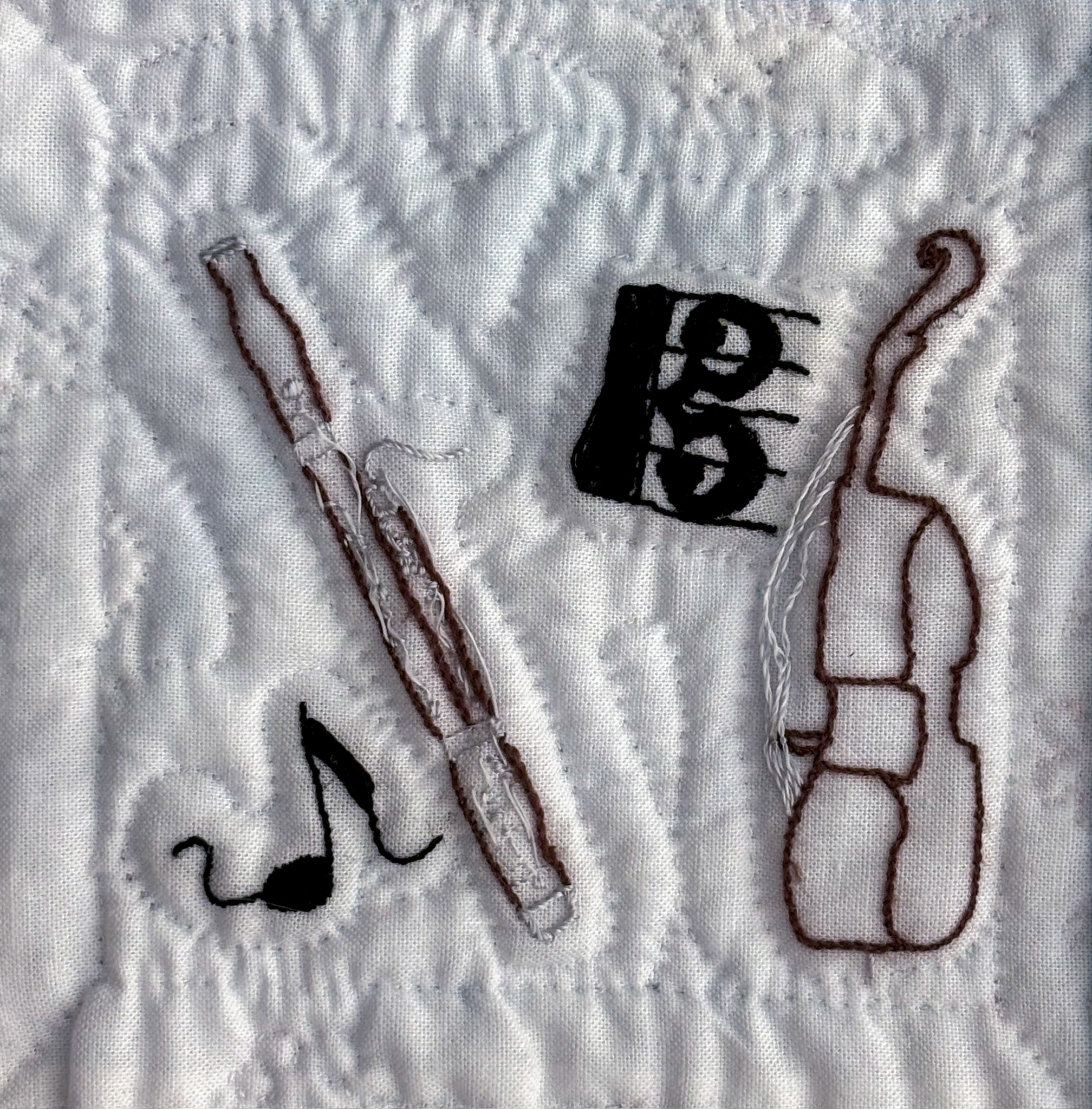 Bassoon and bass outlined in brown with silver accents embroidered on white cotton. Amongst them are a trailing eighth note and an alto clef filled in black.